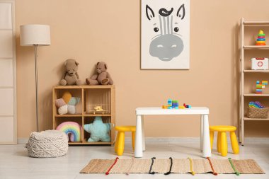 Çocuk odasının içinde lamba, raflarda bir sürü oyuncak ve bej duvarın yanında oyun masası.