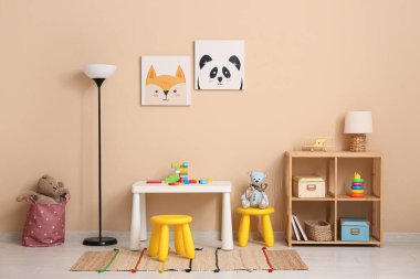 Çocuk odasının içinde oyun masası, lambalar ve bej duvarın yanındaki raflarda bir sürü oyuncak var.