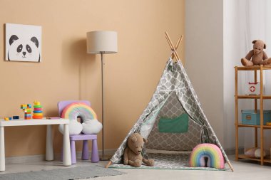 Oyun çadırı, lamba, masa ve bej duvarın yanındaki raflarda bir sürü oyuncakla birlikte çocuk odasının içinde.