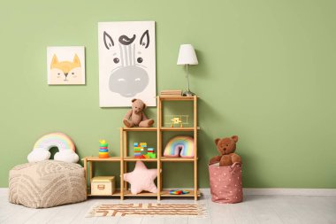 İçinde puf, lamba, raf ünitesinde oyuncaklar ve yeşil duvarda resimler olan çocuk odasının içi.