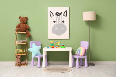 Çocuk odasının içinde lamba, oyun masası ve yeşil duvarın yanındaki raflarda bir sürü oyuncak var.