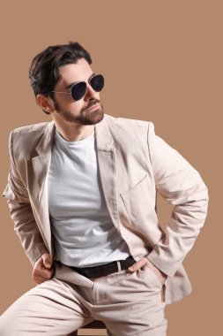 Açık renk takım elbiseli, güneş gözlüklü, bej arka planda, taburede oturan yakışıklı bir genç.