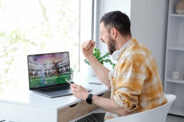 Evdeki spor bahsini kazandıktan sonra cep telefonu ve dizüstü bilgisayarı olan mutlu genç bir adam.