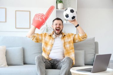 Evdeki kanepede oturan futbol topu ve köpük parmaklı mutlu genç adam. Spor bahsi konsepti