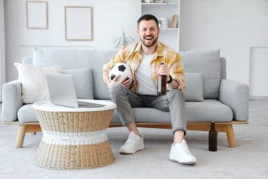 Futbol topu ve birayla evdeki kanepede oturan mutlu genç adam. Spor bahsi konsepti
