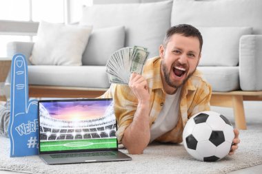 Evde yerde yatan futbol topu ve dizüstü bilgisayarı olan mutlu genç bir adam. Spor bahsi konsepti