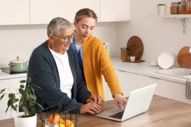 Mutfakta dizüstü bilgisayar kullanan yaşlı bir kadınla genç bakıcı.