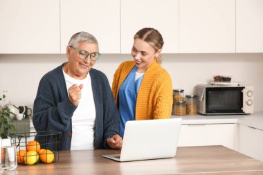 Mutfakta dizüstü bilgisayar kullanan yaşlı bir kadınla genç bakıcı.