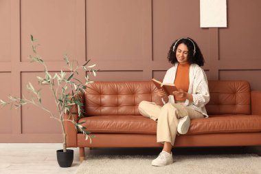 Kulaklıklı genç Afro-Amerikalı kadın evdeki kahverengi duvarın yanındaki kanepede sesli kitap dinliyor.