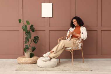 Genç Afro-Amerikalı kadın evdeki kahverengi duvarın yanındaki koltukta cep telefonu kullanıyor.