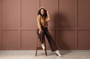 Şapkalı şık bir Afro-Amerikalı kadın kahverengi duvarın yanındaki taburede oturuyor.