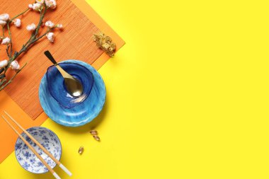 Bamboo mat with bowls, chopsticks and sakura branch on yellow background. Chinese table setting