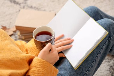 Elinde kahve fincanı olan kadın evde halıda kitap okuyor, yakın plan.