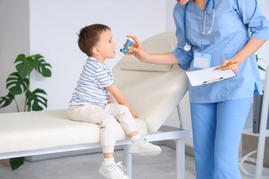 Kadın çocuk doktoru klinikteki kanepede küçük şirin bir çocuğa astım spreyi veriyor.