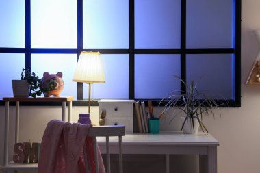 Geceleri pencerenin yanında parıldayan lamba, kitap ve çiçek bulunan şık çocuk odasının içinde.