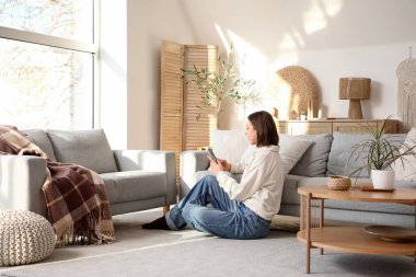 Güzel genç bir kadın büyük pencereli şık bir oturma odasında yerde cep telefonu kullanıyor.