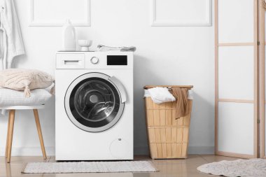 Çamaşır sepetli ve yastıklı modern çamaşır makinesi banyodaki beyaz duvarın yanındaki taburede.