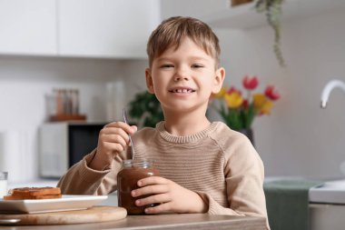 Tatlı çikolata ezmesi kavanozu ve lezzetli tostlarıyla mutfaktaki masada oturan sevimli küçük çocuk.