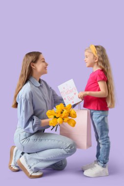Annesini anneler gününde sarı lalelerle, hediye çantasıyla ve eflatun arkaplanlı kartpostalla selamlayan tatlı küçük bir kız.