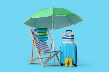Şemsiyeli, valizli çizgili sandalye, plaj çantası ve mavi arka planda parmak arası terlikler.