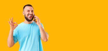 Mutlu genç adam telefonla konuşuyor ve sarı arkaplanda mesaj için yeri olan bir jest gösteriyor.