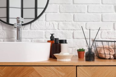 Lavabo kasesi ve banyo aksesuarları beyaz tuğla duvarın yanındaki ahşap dolapta.