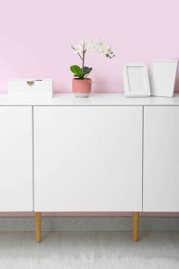 Orkide çiçekli beyaz çekmece ve odada pembe duvarın yanında boş resimler.