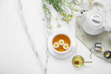 Beyaz mermer arka planda bir fincan sıcak papatya çayı, çaydanlık ve bal.