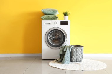 Sarı duvarın yanında battaniye ve çamaşır makinesi olan hasır sepet.