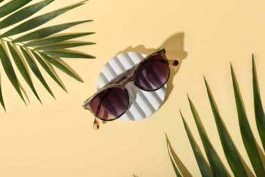 Soluk sarı arka planda şık bir güneş gözlüğü ve palmiye yapraklarıyla dur.