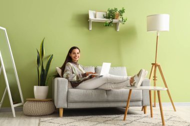 Oturma odasındaki kanepede dizüstü bilgisayarı olan güzel, mutlu genç bir kadın.