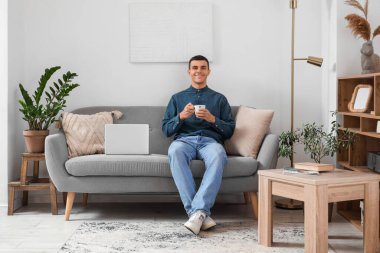 Oturma odasındaki kanepede dinlenen bir fincan kahve ve dizüstü bilgisayarlı yakışıklı genç adam.