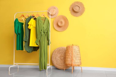 Sarı duvarda şık kadın kıyafetleri ve hasır şapkalar asılı.