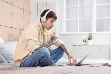 Evde yatağında dizüstü bilgisayar kullanan, kulaklıklı, tek kullanımlık elektronik sigaralı genç yakışıklı bir adam.