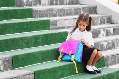 Sırt çantalı küçük kız, okulun yanında oturuyor.