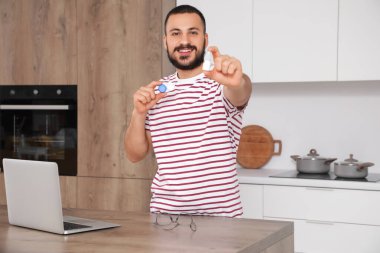 Mutfakta dizüstü bilgisayarın yanında göz damlası ve lens tutan genç bir adam var.