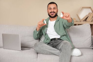 Dizüstü bilgisayarı olan genç adam evdeki kanepede göz damlası tutuyor.