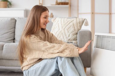 Evde elektrikli konteyner ısıtıcısının yanında ellerini ısıtan genç bir kadın var.