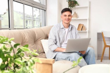 Genç adam evdeki yumuşak koltukta dizüstü bilgisayar kullanıyor.