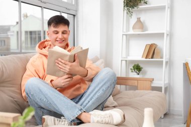 Genç adam evdeki yumuşak kanepede kitap okuyor.
