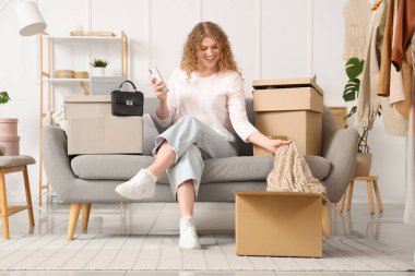 Cep telefonlu genç bir kadın evinde yeni kıyafetlerle birlikte paket kutusunu açıyor.