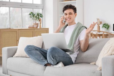 Bilgisayarlı genç adam evdeki koltukta cep telefonuyla konuşuyor.