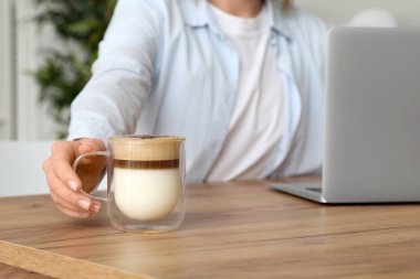 Mutfaktaki masada cam bardak kahve ve modern dizüstü bilgisayarlı genç bir kadın, yakın plan.