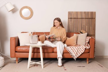 Genç bir kadın dergi okuyor ve oturma odasında kanepede oturuyor.