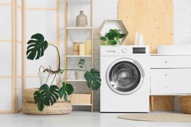 Modern çamaşır makinesi, hasır sepeti, raf ünitesi ve banyo aksesuarları olan şık bir çamaşırhane.