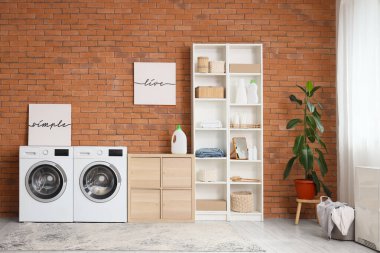 Çamaşır odasının içinde, modern çamaşır makinesi ve kurutma makinesi olan tuğla duvarın yanında.