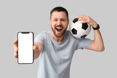 Gri arka planda futbol topu ve cep telefonu olan mutlu genç bir adam. Spor bahsi konsepti