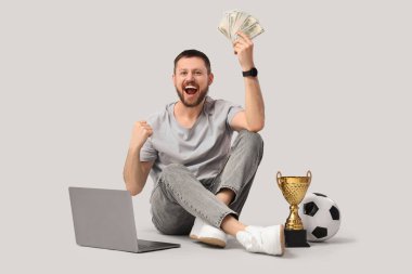 Gri arka planda oturan parası ve bilgisayarı olan mutlu genç adam. Spor bahsi konsepti