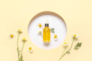 Beyaz arka planda bir şişe kozmetik ürünü ve papatya çiçeği sarı kağıtta yuvarlak delikten görülebiliyor.