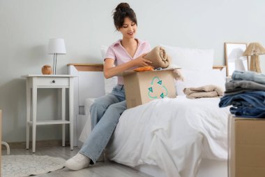 Yatak odasında geri dönüşüm için süveter paketleyen genç bir kadın.
